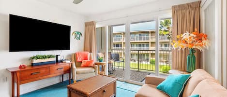 A living room with a flat-screen TV, a ceiling fan, a sofa with teal cushions, a wicker coffee table, and a view of a building and greenery through the glass doors.