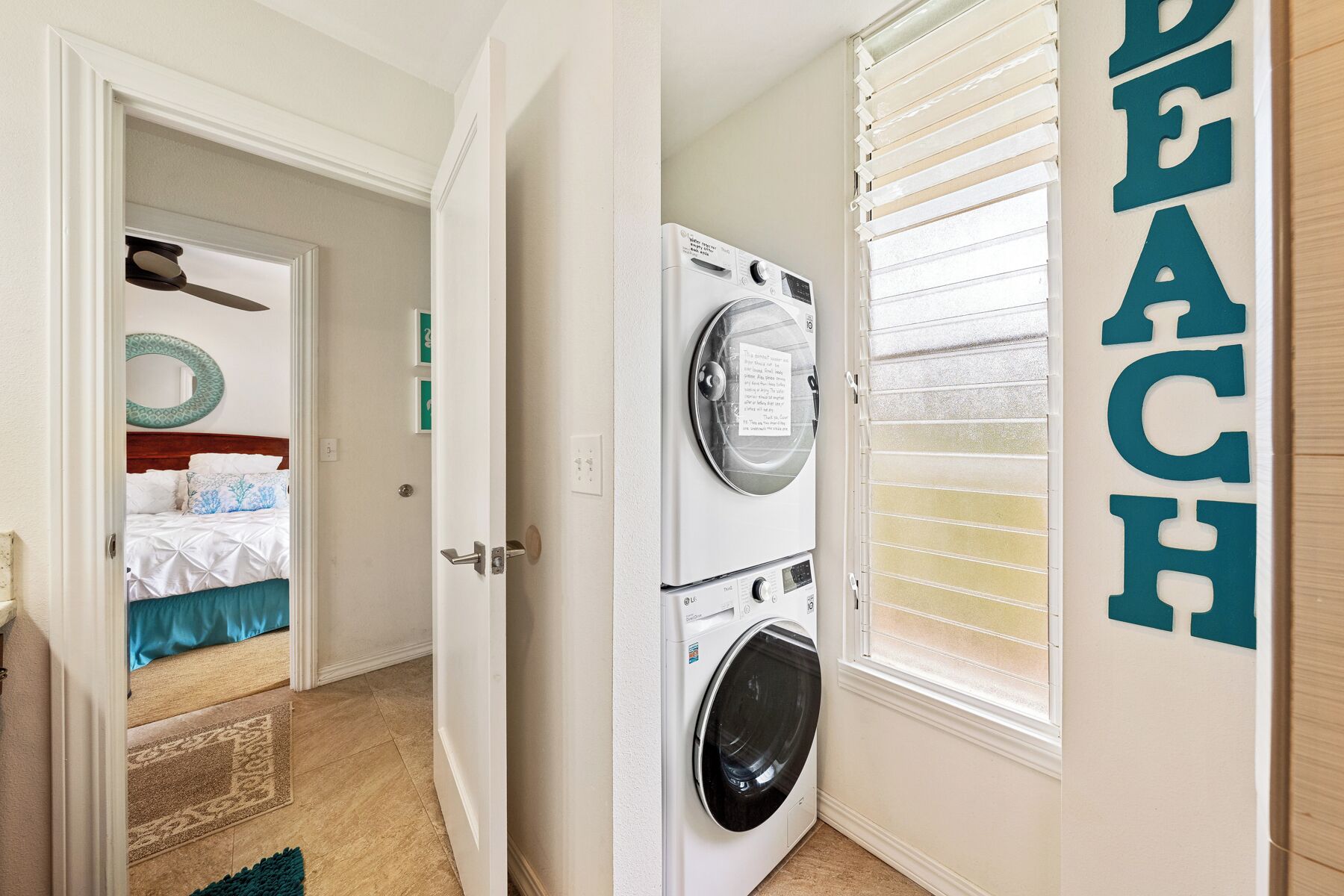 A laundry room with a washing machine and dryer, a door leading to a bedroom with a bed and a circular mirror, and a wall with the word "BEACH" in teal letters.