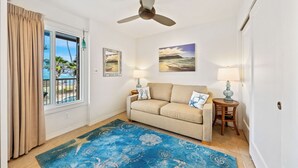 Ein Wohnzimmer mit einem beigefarbenen Sofa, zwei Wandlampen, einem Deckenventilator, einem blauen Teppich mit Muster und einem großen Fenster mit Vorhängen.