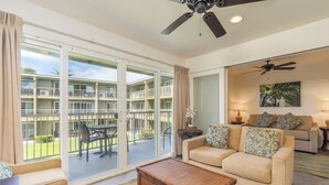 A spacious living area with a sliding glass door leading to a balcony, a beige sofa with patterned cushions, a wicker coffee table, and a ceiling fan. - Privacy, Ocean View! Modern Kitchen, WiFi, Flat Screen, Lanai–Kauai Kailani K212 (Kapa'a)