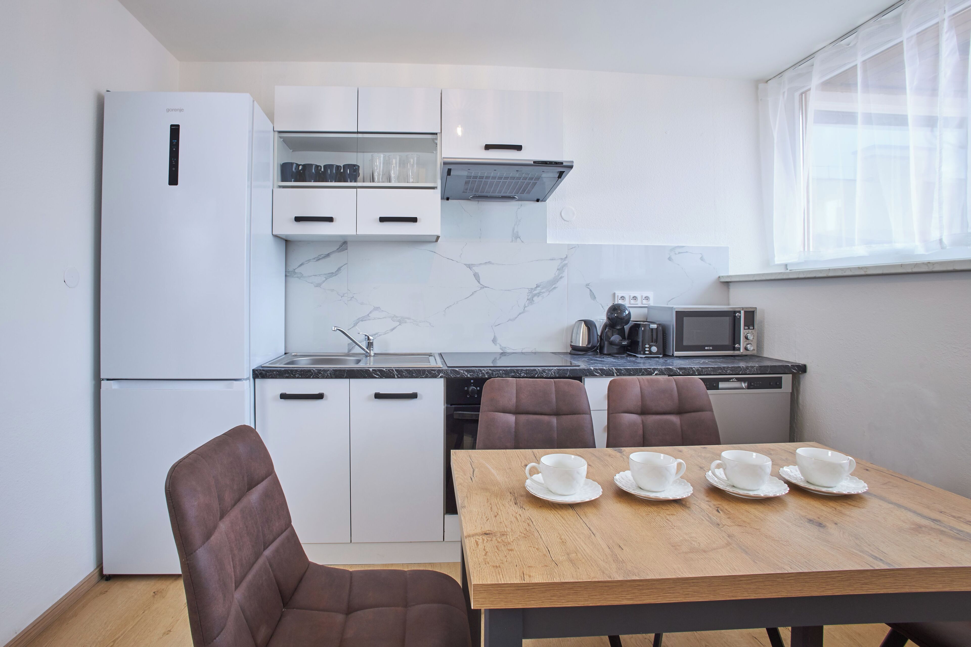 A modern kitchen with white cabinets, a refrigerator, a microwave, and a coffee maker. There is a dining table with four chairs and four white cups.