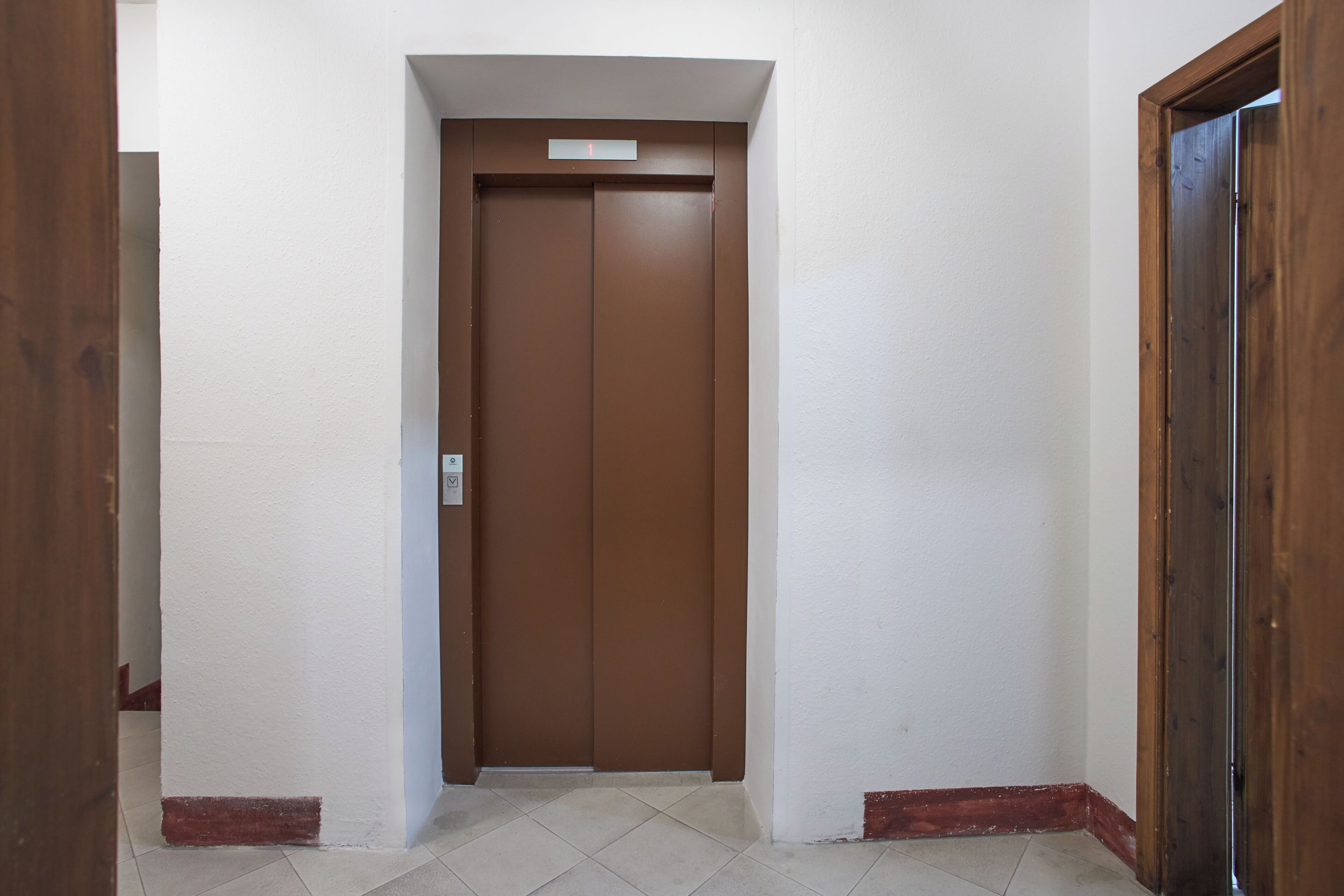 A narrow hallway with a brown elevator door, white walls, and a tiled floor.