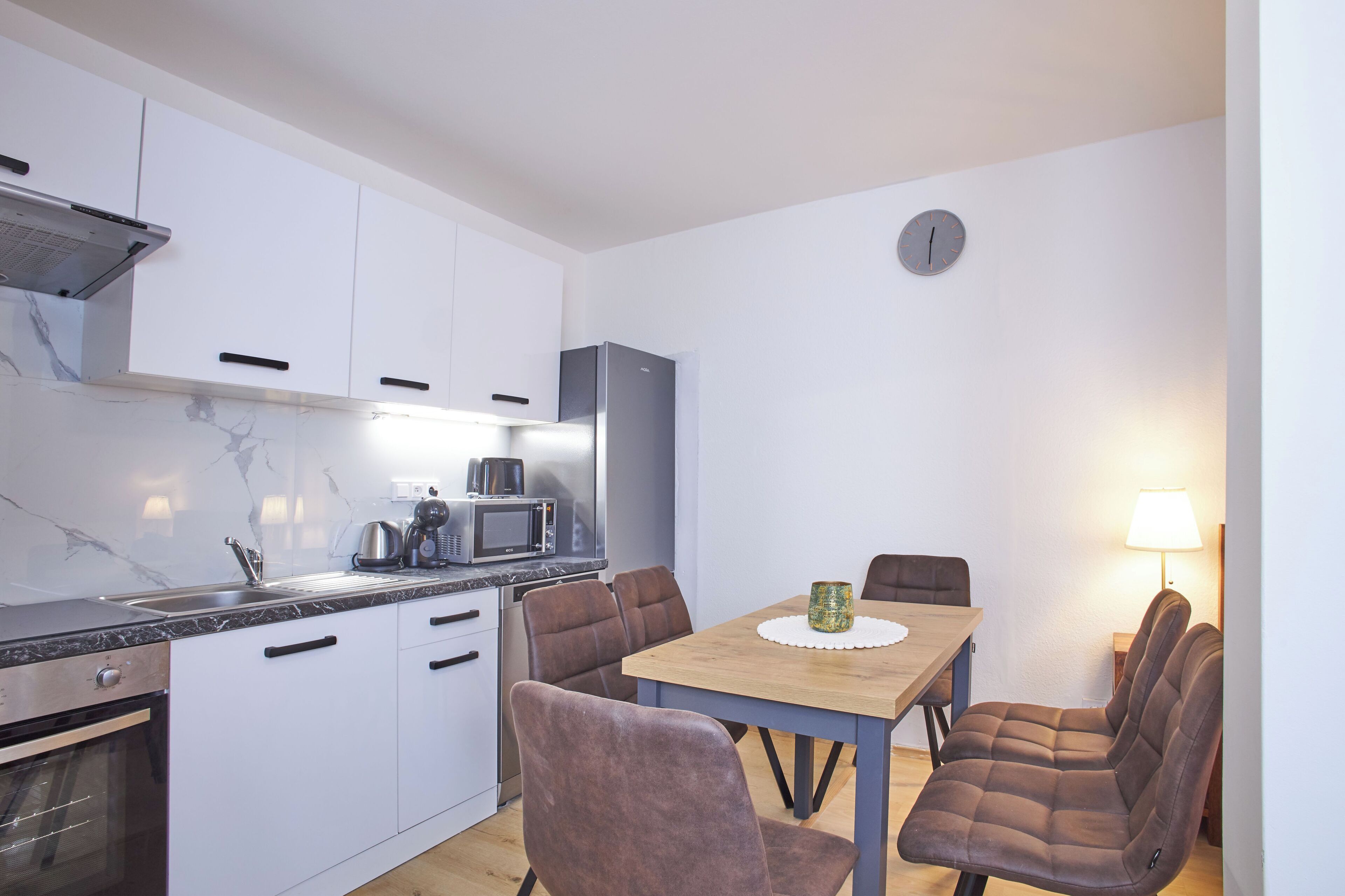 Cocina compacta con armarios blancos, microondas y cafetera. Cuenta con una mesa de comedor con cuatro sillas y una lámpara pequeña.