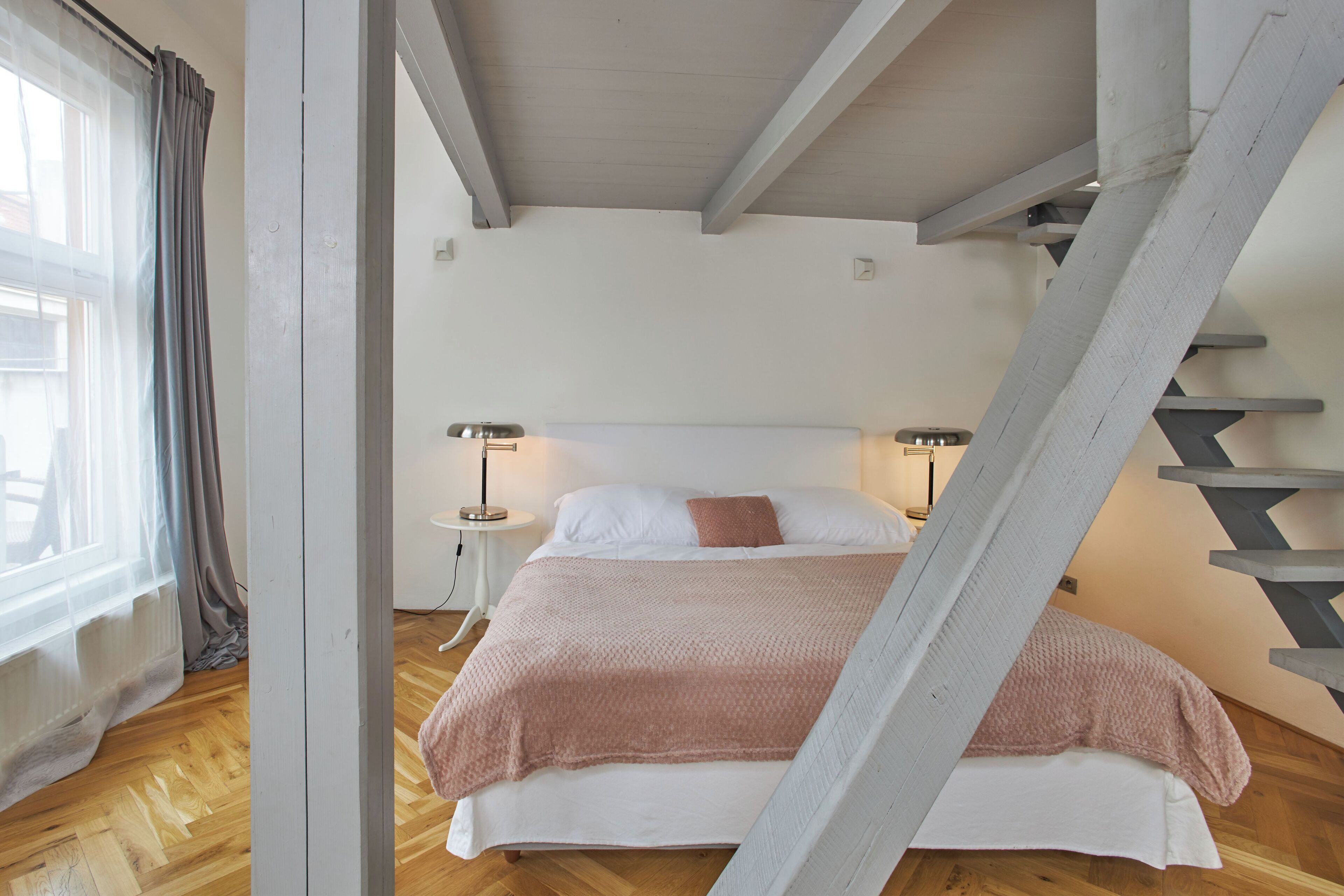A bedroom with a bed, two bedside lamps, a window with curtains, and a wooden staircase.