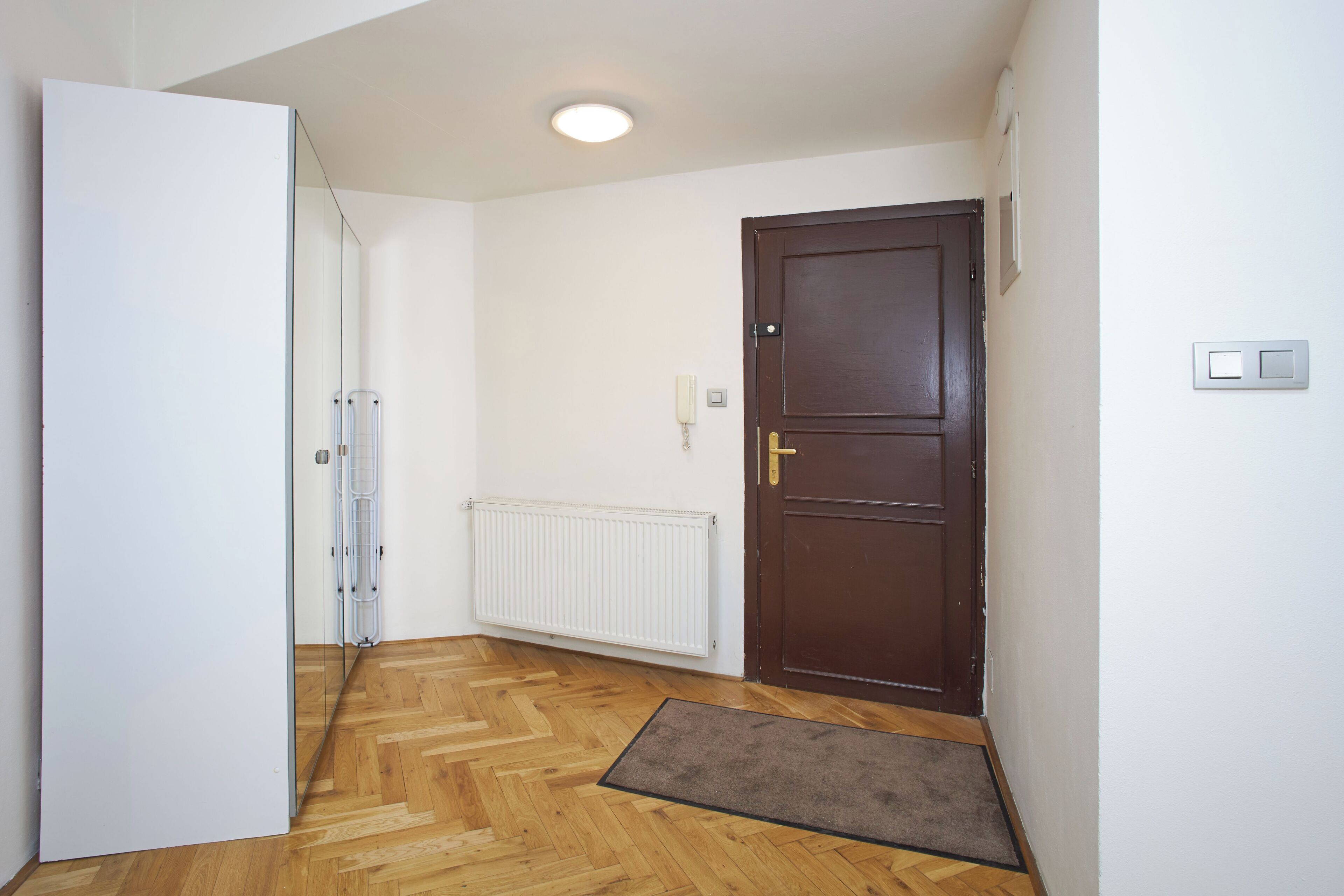 Un pasillo con suelo de madera, un radiador, una puerta marrón y una pared blanca.