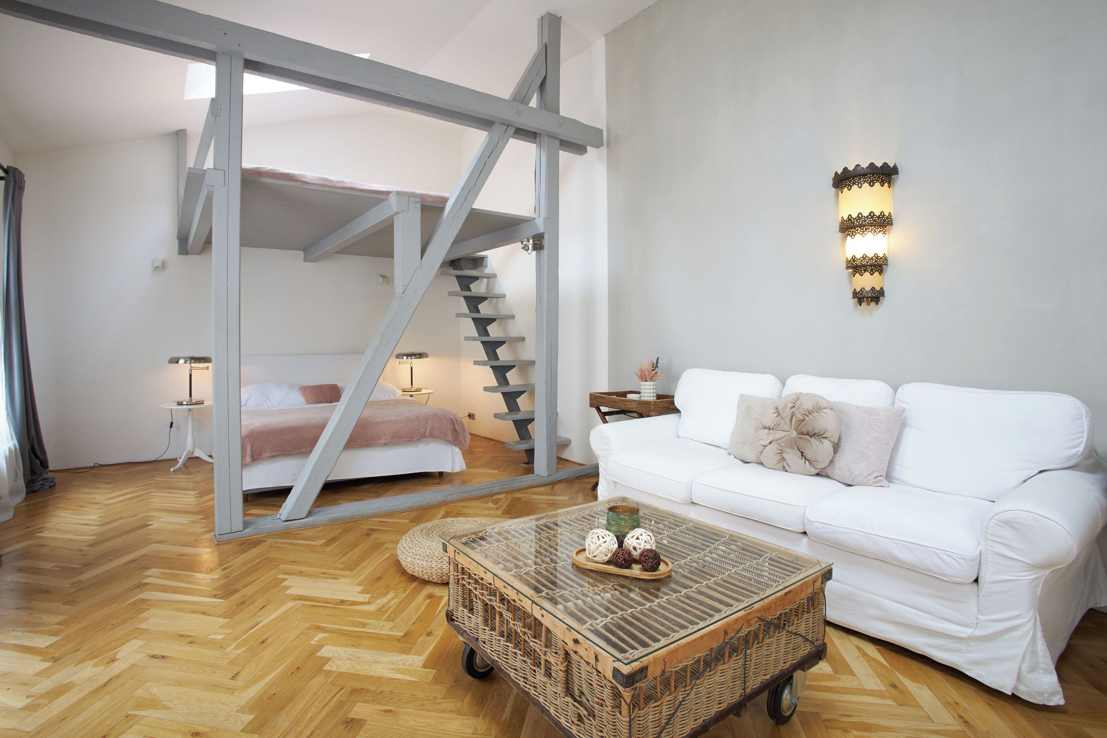 A room with a white sofa, a glass coffee table with decorative items, a wicker side table, a white bed with a pink blanket, and a staircase with a grey railing.