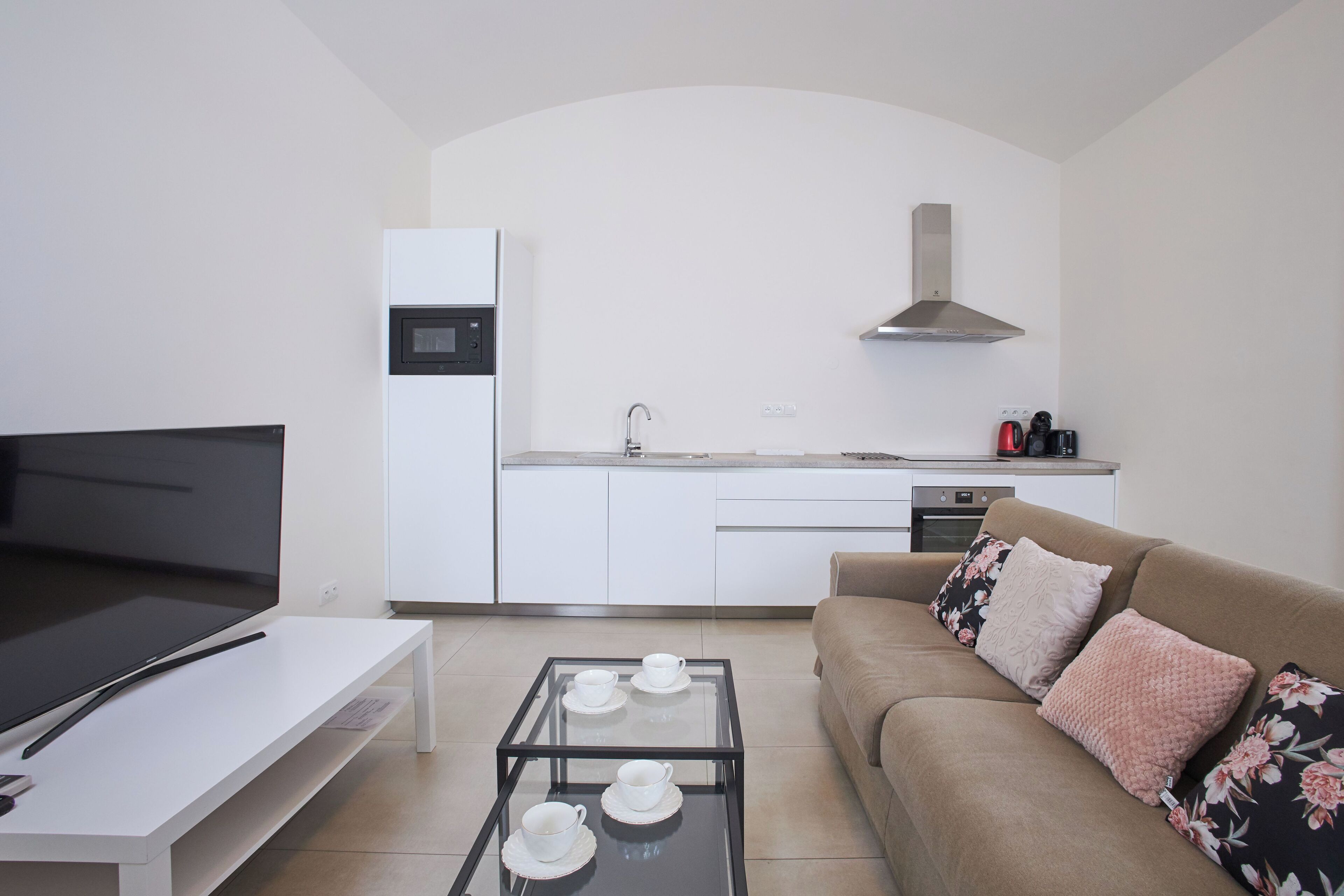 Una sala moderna con un televisor de pantalla plana, una mesa de centro de vidrio con tazas, un sofá con cojines estampados y una cocina al fondo.
