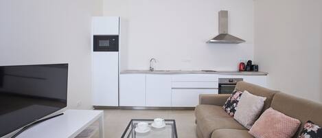 Una sala moderna con un televisor de pantalla plana, una mesa de centro de vidrio con tazas, un sofá con cojines estampados y una cocina al fondo.