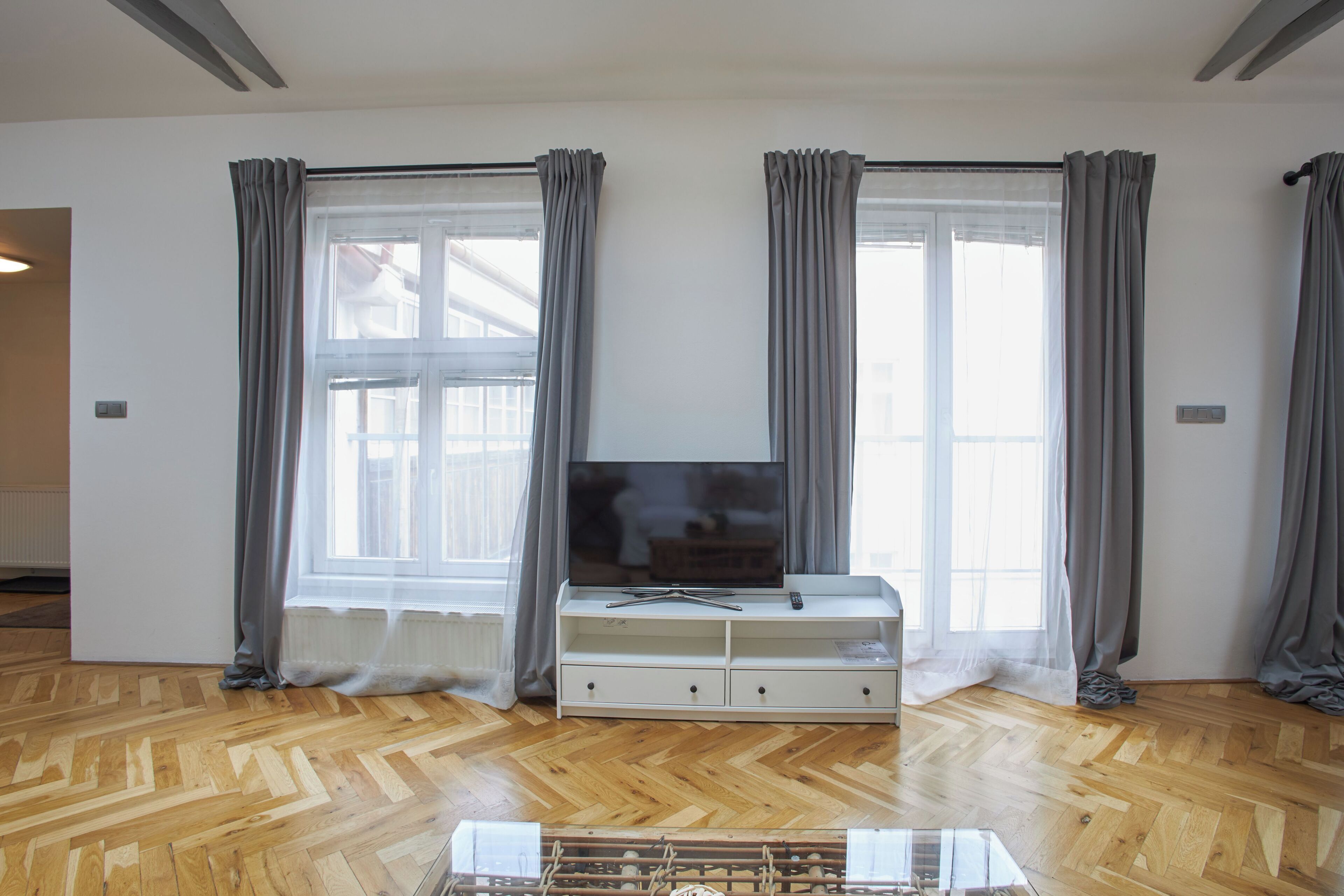 A room with a wooden parquet floor, a white entertainment unit with a flat-screen TV, and grey curtains on the windows.