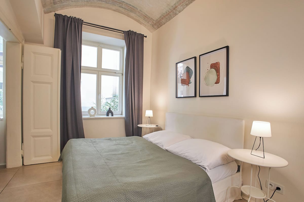 A neatly arranged bedroom with a bed, a bedside table with a lamp, a window with curtains, and two framed pictures on the wall.