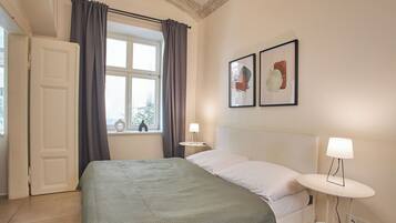 A neatly arranged bedroom with a bed, a bedside table with a lamp, a window with curtains, and two framed pictures on the wall.