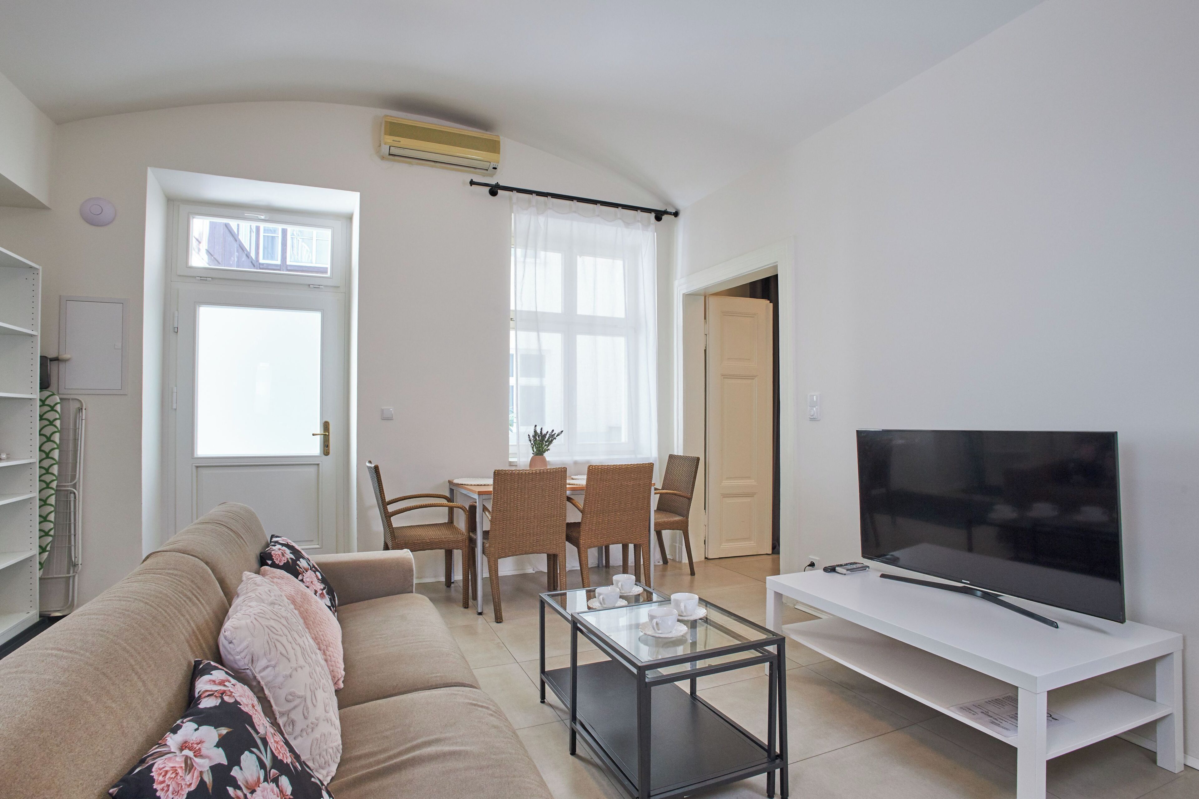A living room with a sofa, a coffee table, a TV on a stand, a dining area with chairs and a table, and a door leading to another room.