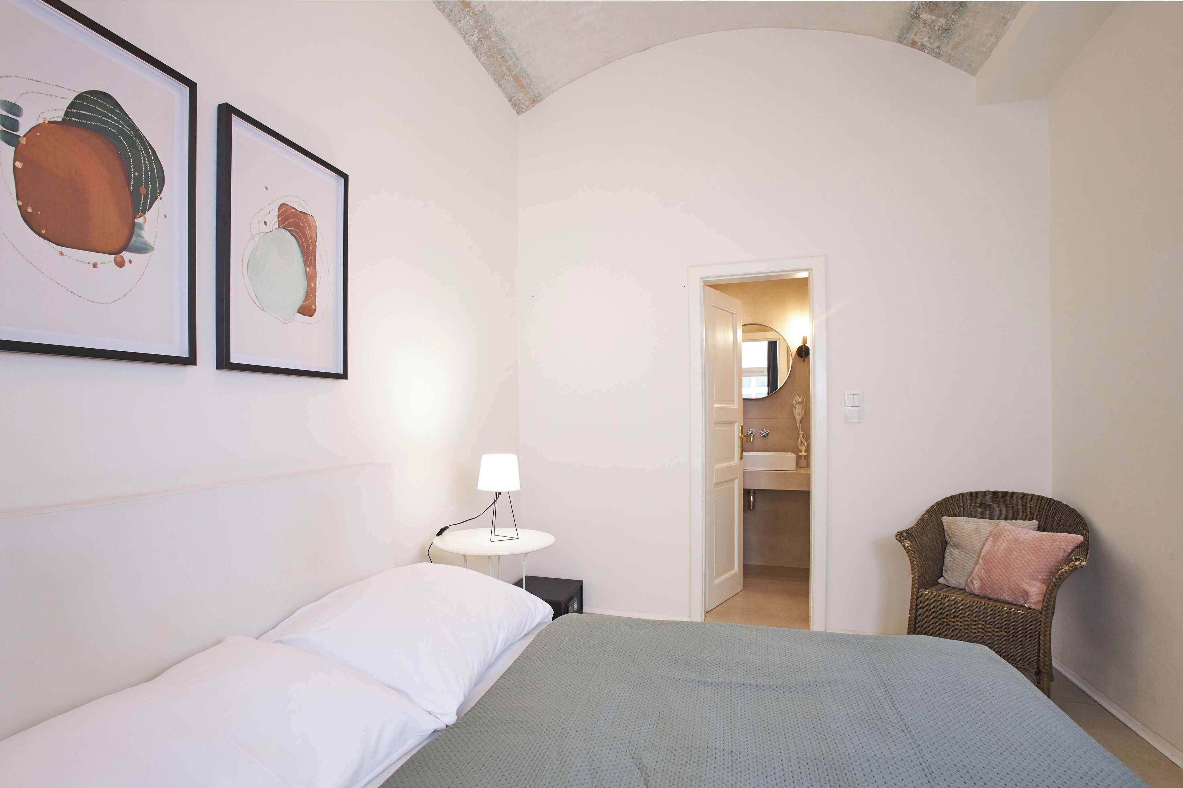 A hotel room with a bed, a chair, a small table with a lamp, and two framed artworks on the wall.