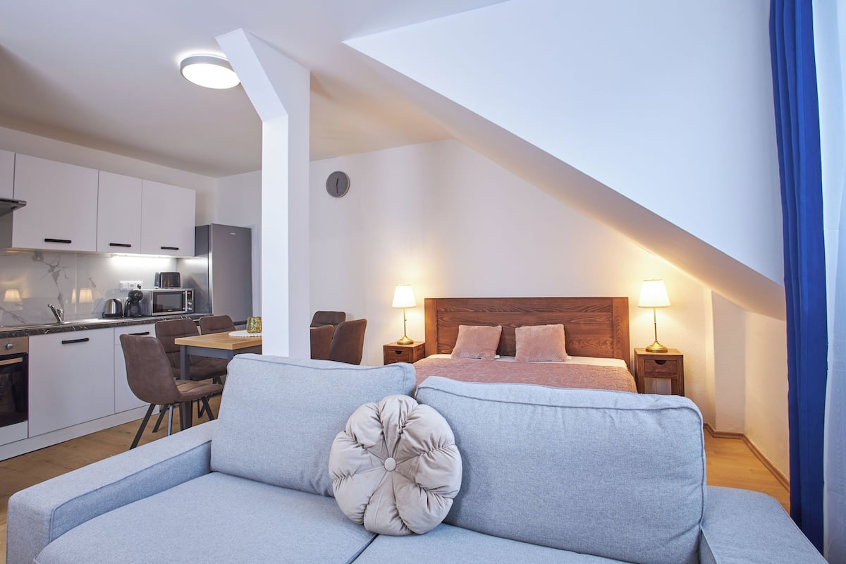 A modern bedroom with a large bed, a sitting area with a sofa, and a kitchenette in the background.