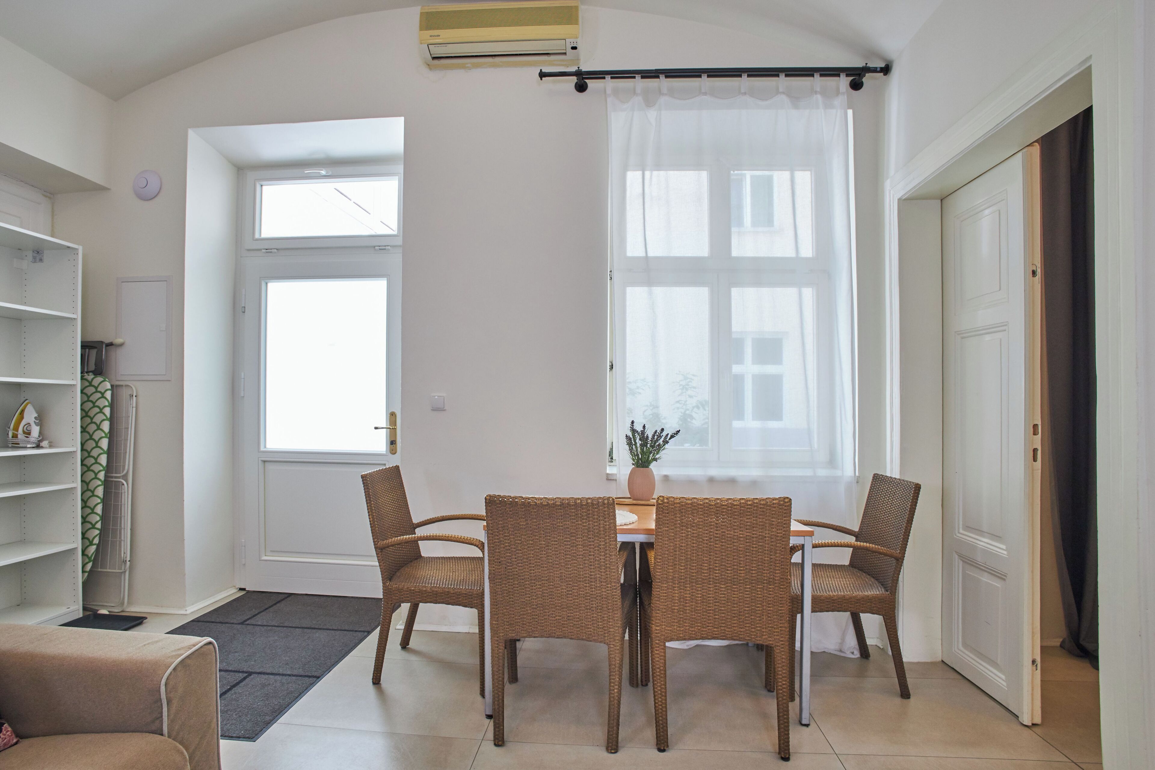 A room with a dining table, four wicker chairs, a potted plant, a radiator, and a large window with a curtain.
