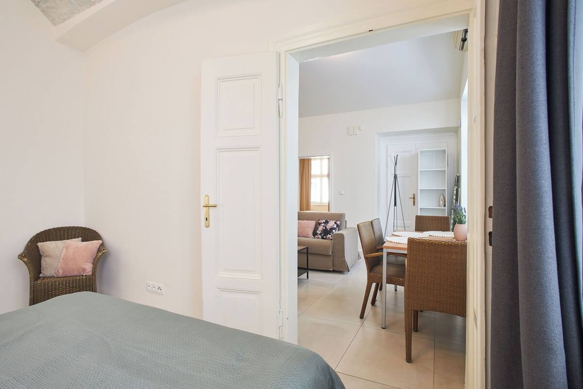A bedroom with a bed, a wicker chair, and a door leading to a living area with a sofa and a dining table.