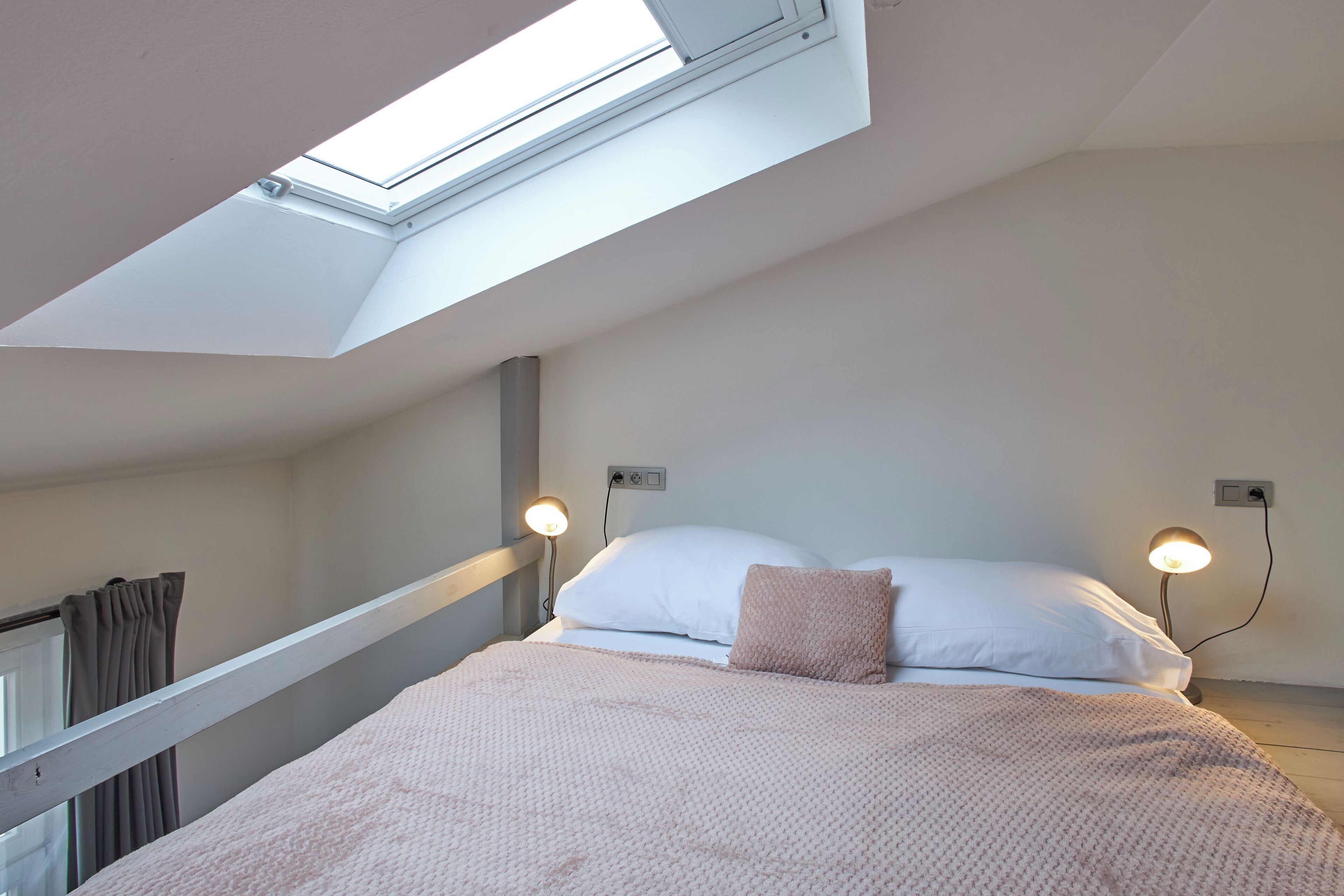 Una camera da letto con un velux, due lampade ai comodini, un letto con un cuscino rosa e una finestra con tende.