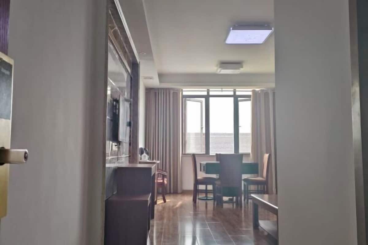 A hotel room with a dining area, a large window with curtains, and a ceiling light.
