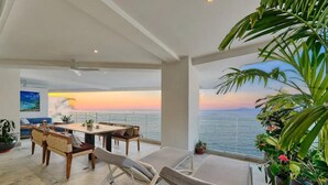 Ein geräumiges Wohnzimmer mit einem großen Esstisch, bequemen Sitzgelegenheiten und Blick auf den Sonnenuntergang über dem Meer.