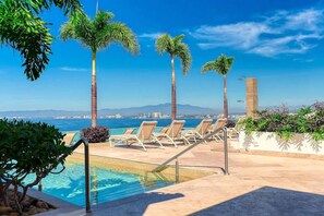 Ein Poolbereich mit Liegestühlen, Palmen und Blick auf das Meer und die Stadtlandschaft.