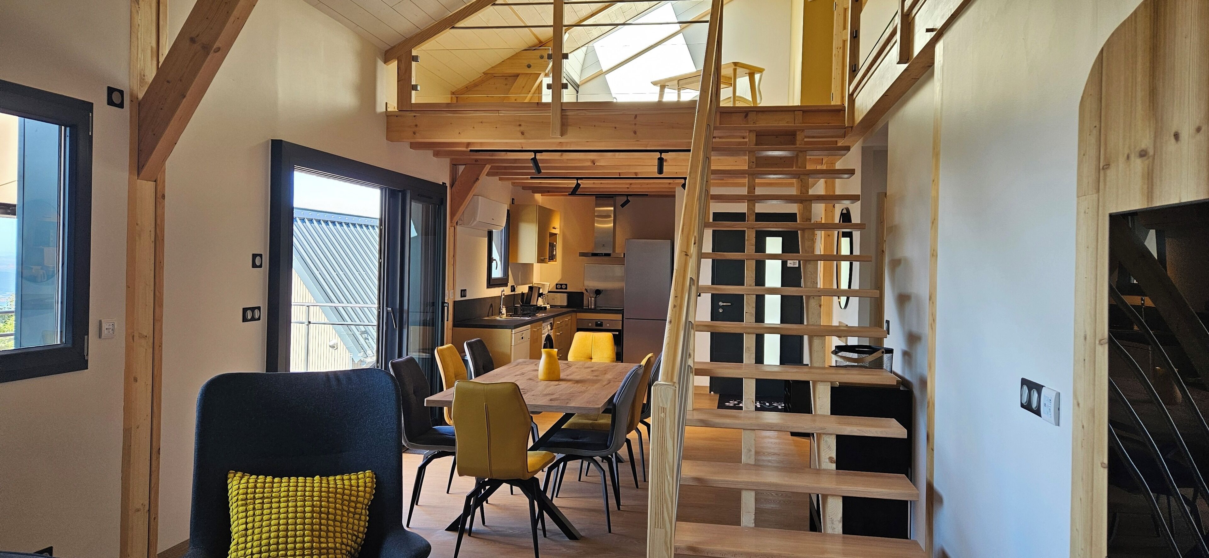 Un interior moderno con una escalera de madera que conduce a un altillo, un comedor con sillas amarillas y una cocina al fondo.