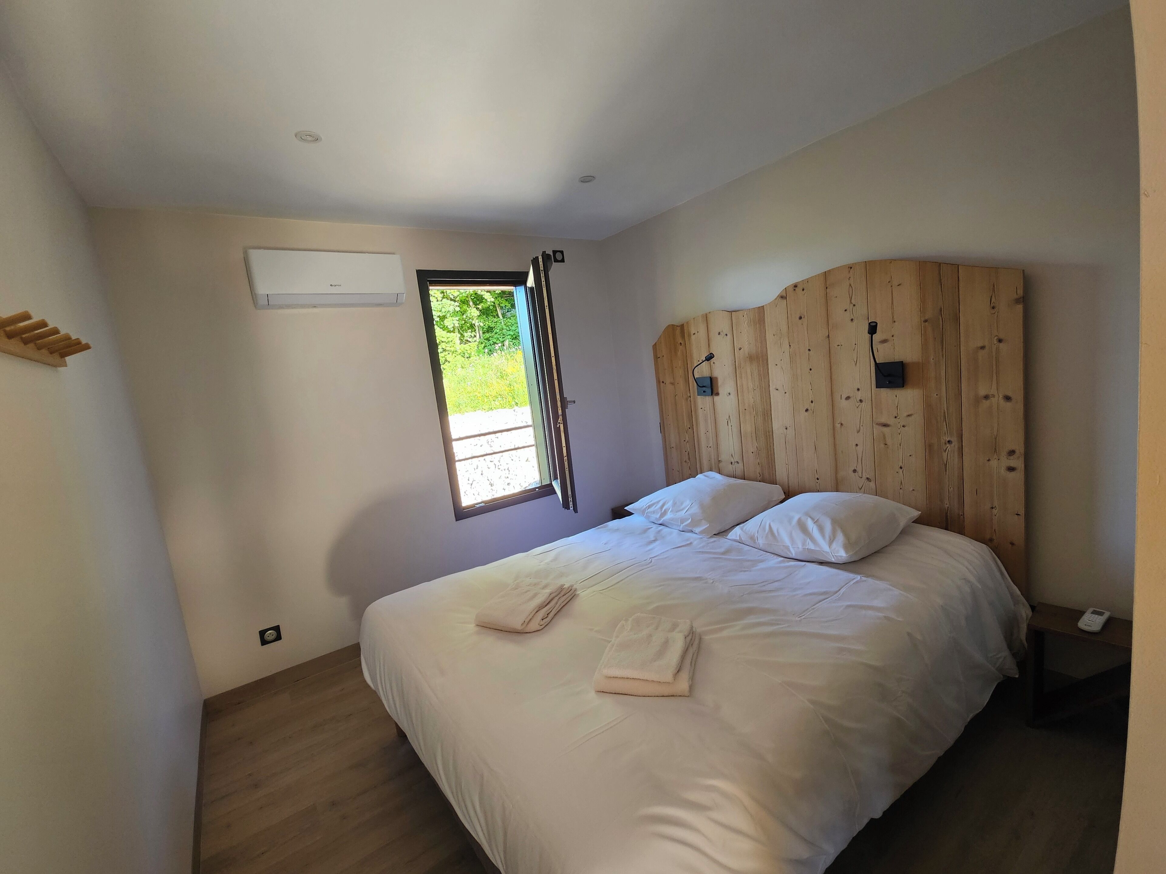 Quarto com cabeceira de madeira, uma janela grande e uma cama bem arrumada com duas almofadas e uma toalha dobrada.