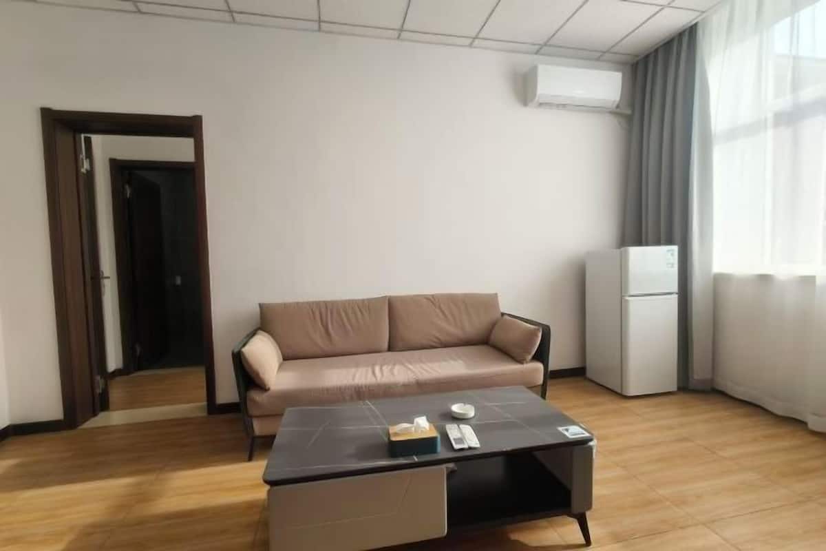 Una sala de estar moderna con un sofá color beige, una mesa de centro negra con una caja de pañuelos y un control remoto, un refrigerador blanco y una puerta que da a otra habitación.