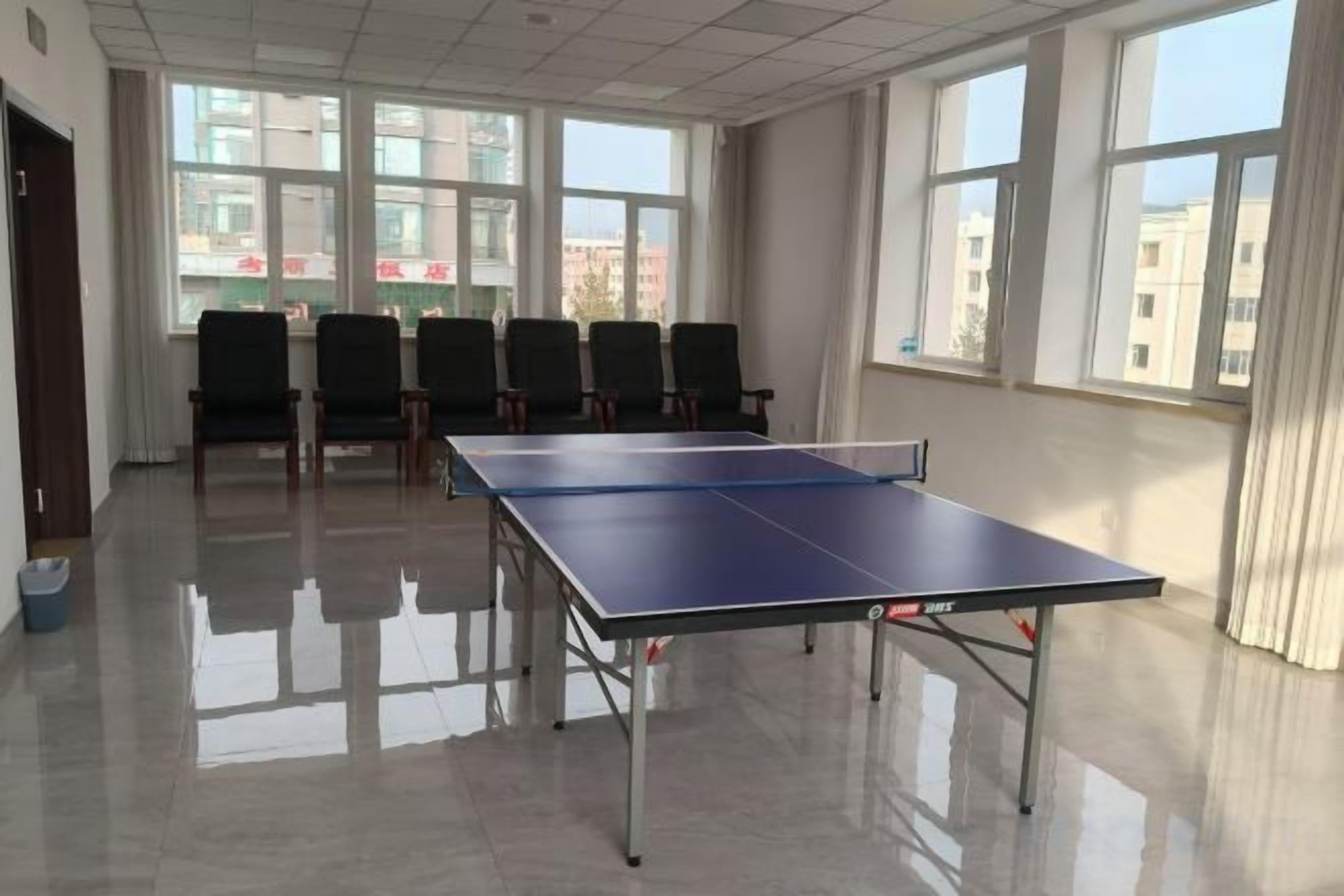 A room with a ping pong table, chairs, and large windows.