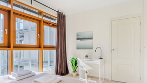 A bedroom with a bed, a desk, a potted plant, a window with curtains, and a door. - P&O Apartments Arkadia 1 (Warsaw)