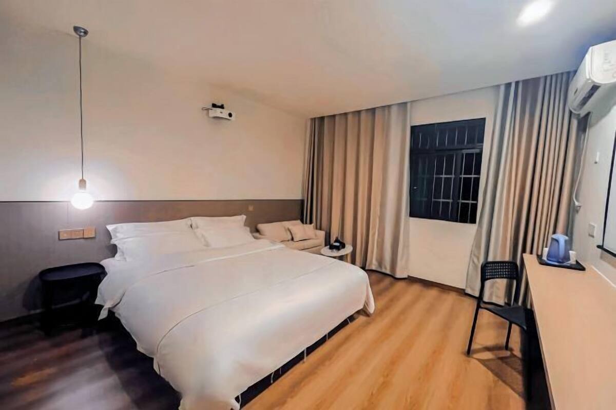 Quarto de hotel com uma cama grande, uma mesa, uma cadeira, uma mesinha, um abajur, uma janela com cortinas e um ar condicionado.