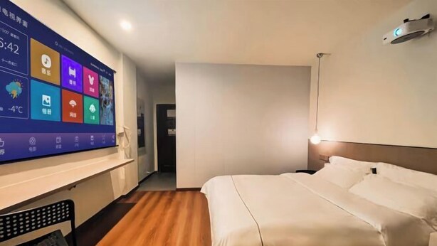 A hotel room with a bed, a desk, and a large screen displaying room information.