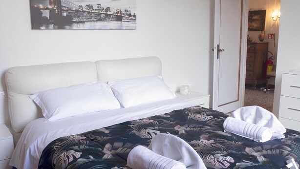 A neatly made bed with white linens, a floral-patterned bedspread, and two rolled towels. A cityscape painting hangs above the bed. - Ponchielli 8 Vintage Flat (Florence)