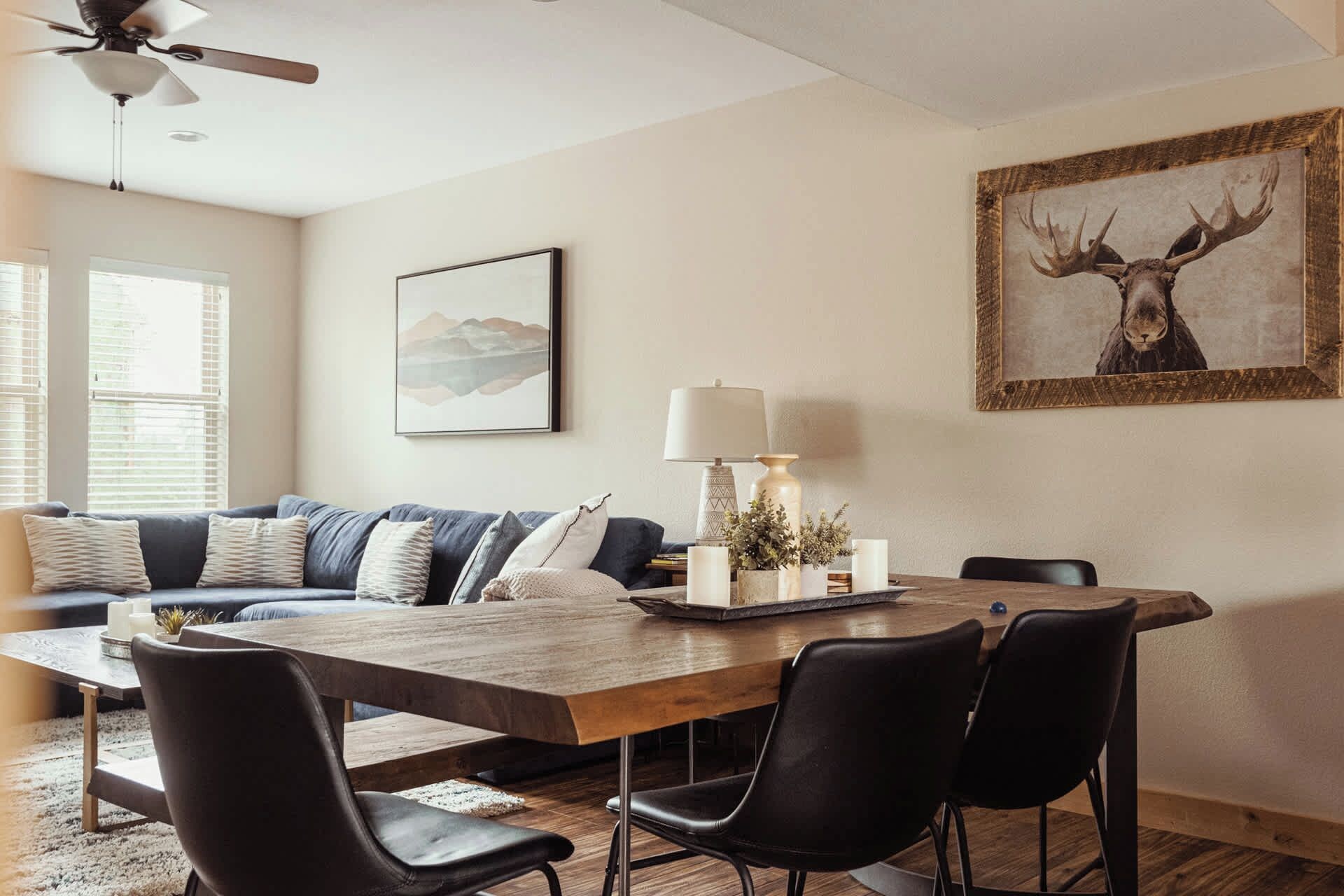 A modern living room with a wooden dining table, black chairs, a sofa with patterned cushions, a ceiling fan, and a framed deer picture on the wall.