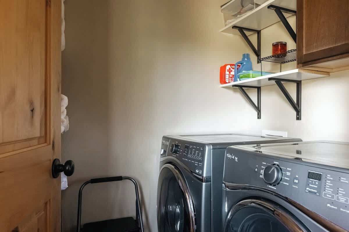Een kleine wasruimte met een voorlader wasmachine, een metalen plank met schoonmaakspullen, en een deur met een handdoekdispenser.