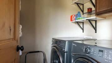 Een kleine wasruimte met een voorlader wasmachine, een metalen plank met schoonmaakspullen, en een deur met een handdoekdispenser.