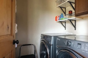 Pequeña lavandería con lavadora de carga frontal, un estante metálico encima que contiene productos de limpieza, y una puerta con dispensador de toallas.
