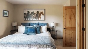 Un dormitorio con una cama, dos mesitas de noche, un cuadro colgado en la pared de caballos, una puerta de madera y un ventilador de techo.