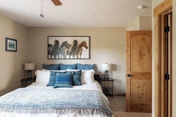 A bedroom with a bed, two bedside tables, a wall-mounted painting of horses, a wooden door, and a ceiling fan.