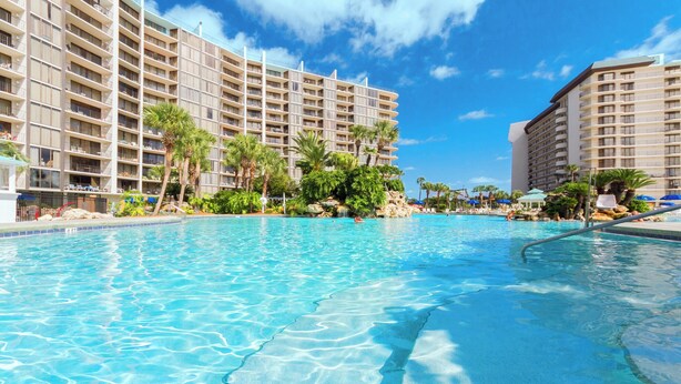 A large swimming pool with clear blue water, surrounded by palm trees and a multi-story hotel building.
