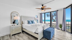 A bedroom with a bed, bedside tables, a ceiling fan, a large window with a view of the beach, and a balcony.