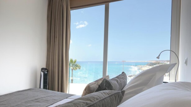 A bedroom with a large window overlooking a beach and ocean.