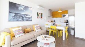 A modern living room with a sofa, a coffee table, and a dining area with yellow chairs. There is a kitchen in the background with yellow cabinets and a microwave.