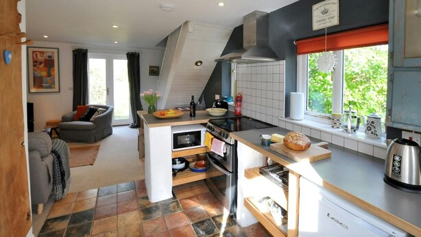 A modern kitchen with a central island, a stove, and a sink. There is a dining area with a couch and a window with a view of greenery. - Y Cwtch, Newport - One Bedroom Cottage, Sleeps 2 (Newport)