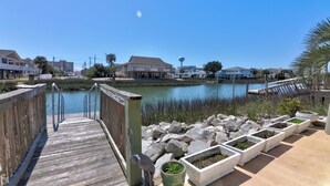 A wooden boardwalk leading to a waterfront area with a clear view of a lake, buildings in the background, and a palm tree on the right.