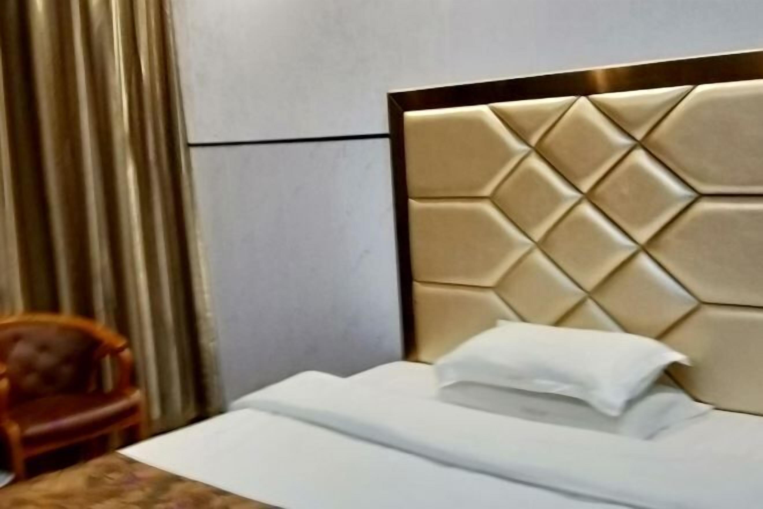 A neatly made bed with a padded headboard, a wooden chair, and a curtain in the background.