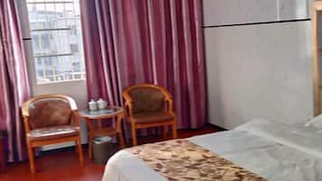 A hotel room with a bed, two wooden chairs, a small round table with a kettle, and a window with bars.