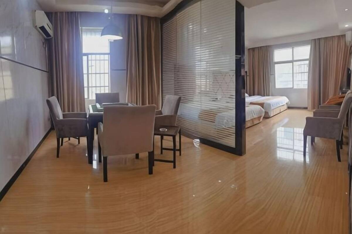 A hotel room with a wooden floor, a dining table with chairs, a sofa, a bed, and a large window with curtains.