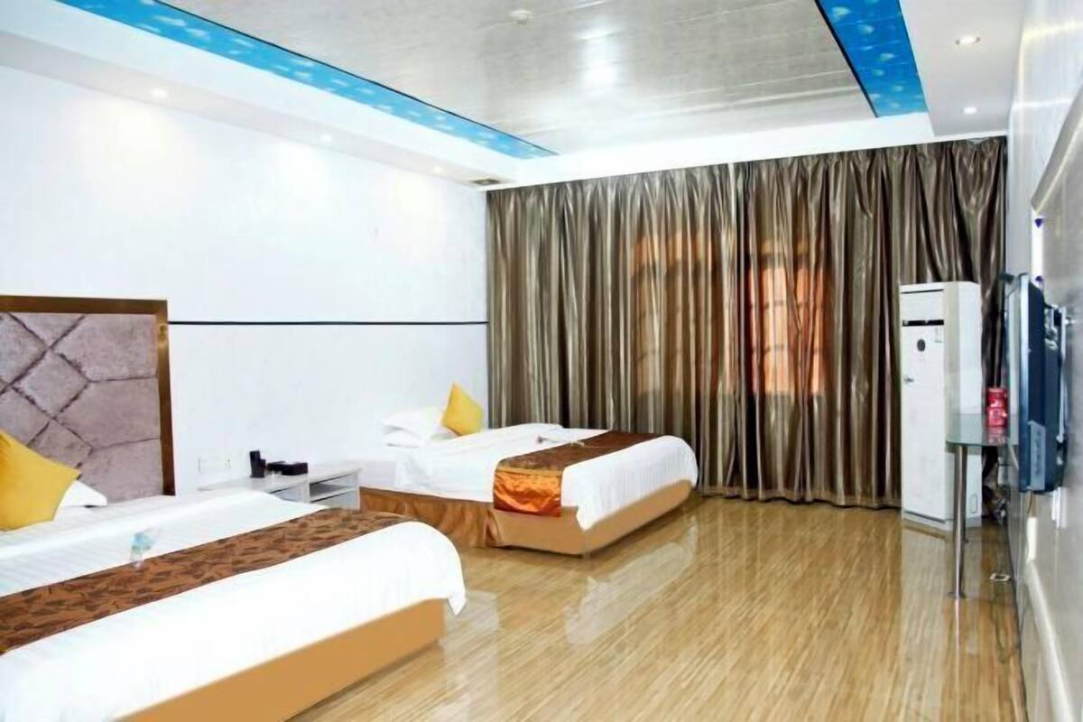 A hotel room with two beds, a flat-screen TV, a minibar, and a large window with curtains.