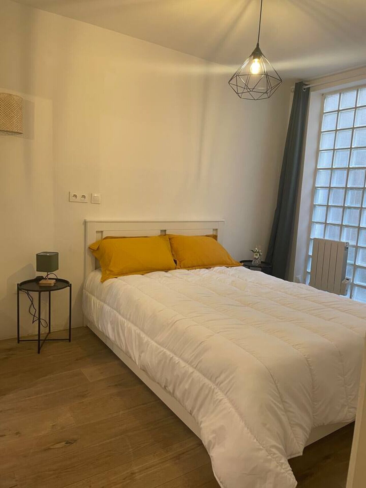 A bedroom with a bed, a bedside table with a lamp, a window with curtains, and a hanging light fixture.