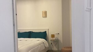 A bedroom with a bed, a bedside lamp, a small round table, and a woven stool. - Luma Oberkampf Apartment / Jean Pierre Timbaud (Paris)